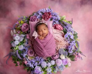 Sleeping newborn wrapped in purple swaddle nestled in wreath of colorful purple and pink flowers.