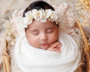 Peaceful newborn baby wrapped in white swaddle wearing floral crown sleeping in wicker basket.