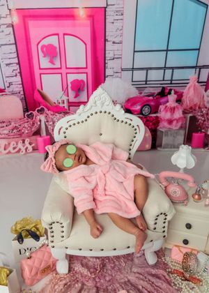 Newborn baby in pink robe with cucumber eye mask sleeping in Barbie dreamhouse spa setting.