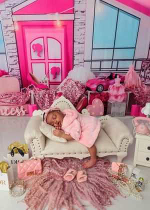 Newborn baby sleeping on white sofa in pink Barbie-themed nursery with luxury accessories.