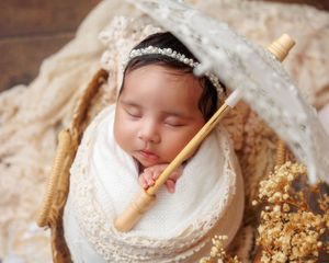 Sleeping newborn swaddled in white wrap inside basket with lace umbrella and dried flowers.