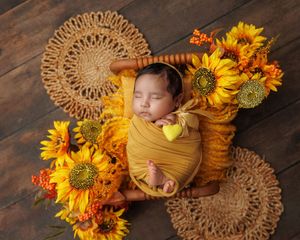 Sleeping newborn wrapped in mustard yellow swaddle surrounded by sunflowers in wooden crate.