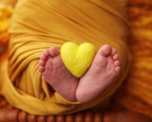 Tiny newborn baby feet holding a yellow felt heart wrapped in golden swaddle cloth.