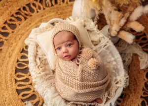 Alert newborn in beige knit wrap and bonnet sitting in wicker basket with boho decor.