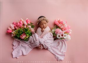 Newborn baby wrapped in white swaddle with floral headband lying between pink tulip bouquets on pink background.