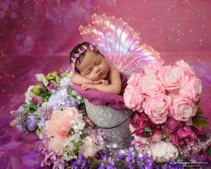 Sleeping newborn with iridescent fairy wings surrounded by pink roses and purple flowers on magenta backdrop.