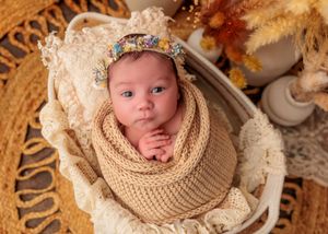 Alert newborn baby wrapped in tan knit swaddle with floral headband lying in white wicker basket.