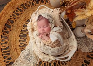 Sleeping newborn in cream lace bonnet and swaddle resting in wicker basket on woven rattan mat.