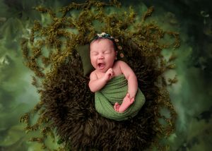 Yawning newborn baby wrapped in green swaddle lying in mossy nest with green leafy background.