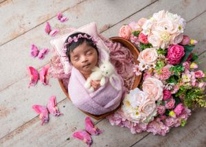 Newborn baby in pink bonnet holding white bunny surrounded by roses and butterflies.