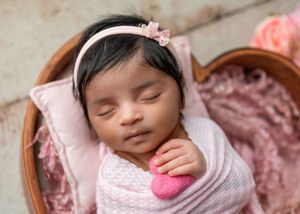 Sleeping newborn baby wrapped in pink holding a felt heart in a wooden bowl.