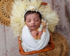 Swaddled newborn baby sleeping in rattan basket holding small deer stuffed animal with floral headband.