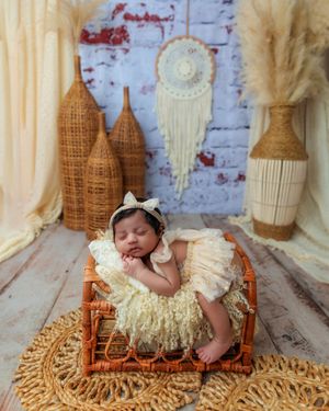 Newborn baby sleeping in rattan crib wearing cream ruffle dress with boho-style backdrop decor.