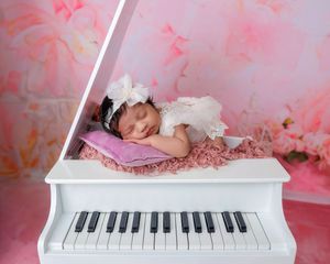 Sleeping newborn baby girl in white outfit resting on miniature white grand piano with pink backdrop.