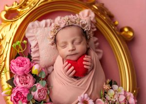 Sleeping newborn in pink wrap holding red heart with floral bonnet in gold frame.