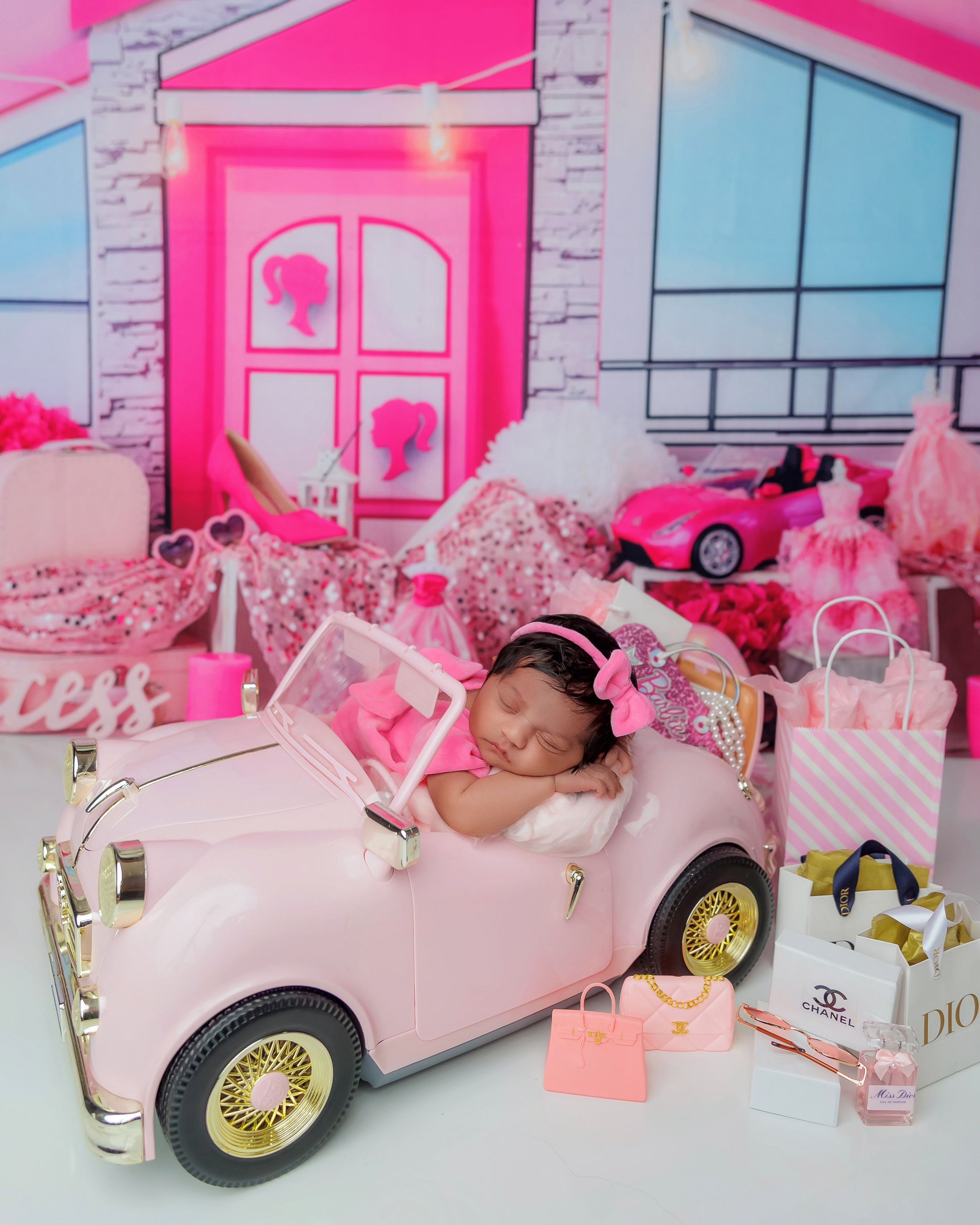 Sleeping newborn baby in pink toy car surrounded by Barbie-themed pink backdrop and accessories.