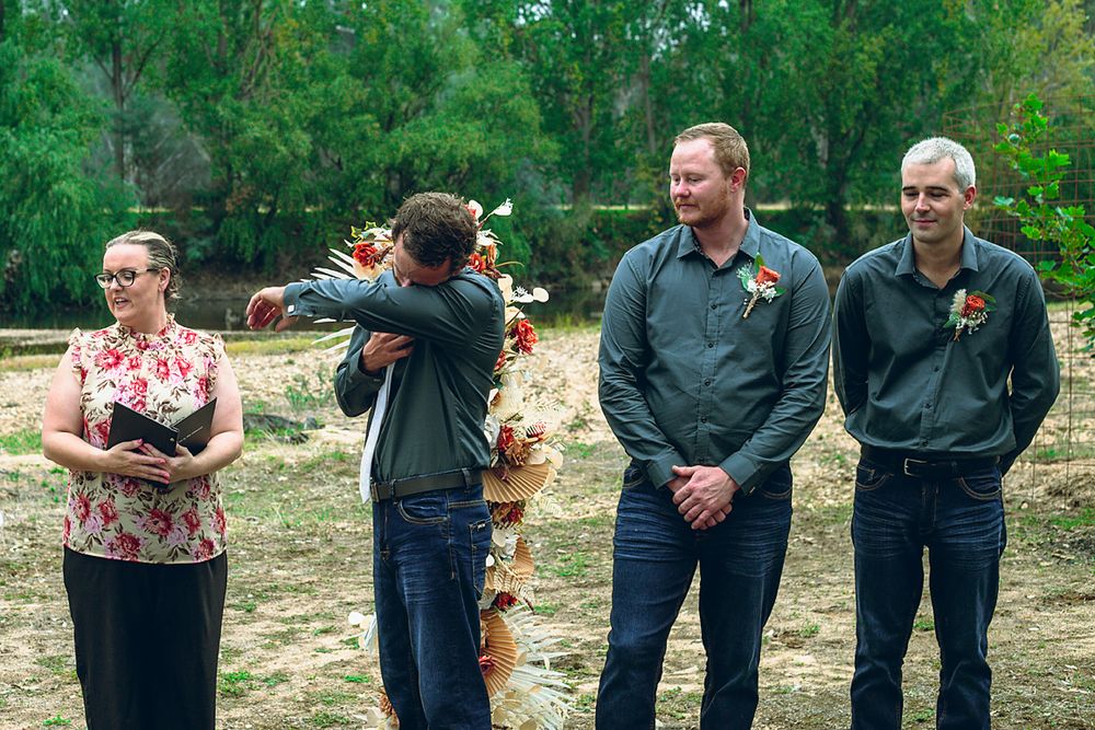 A group of people in dark clothing stand in a line looking down at the ground in a natural outdoor setting.