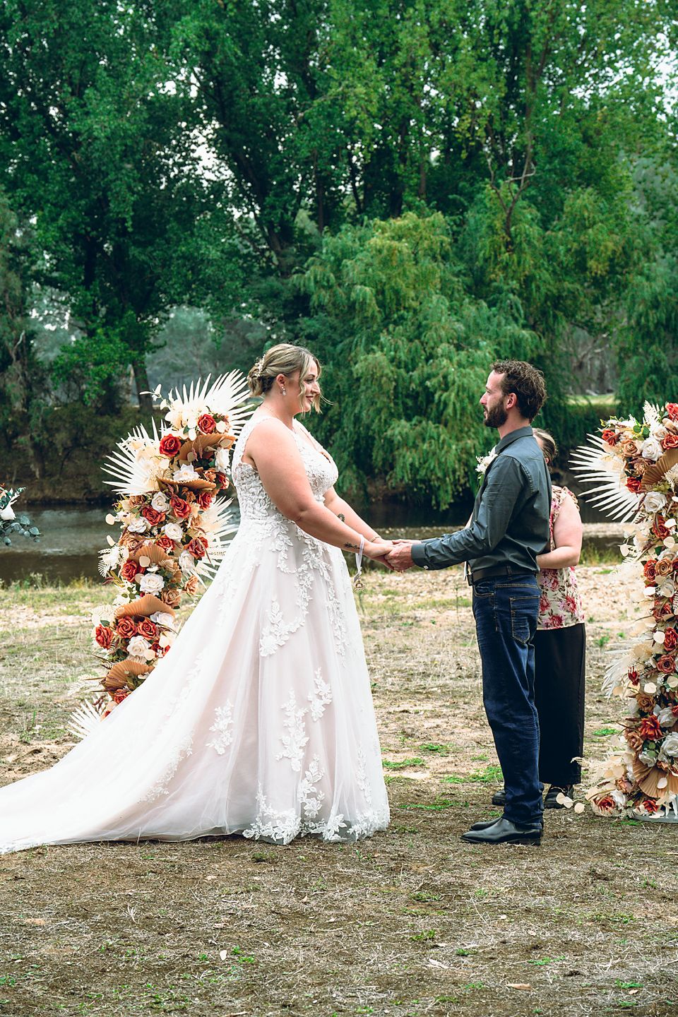 A wedding ceremony takes place outdoors with a floral arch decorated in pink and white blooms against a green backdrop.
