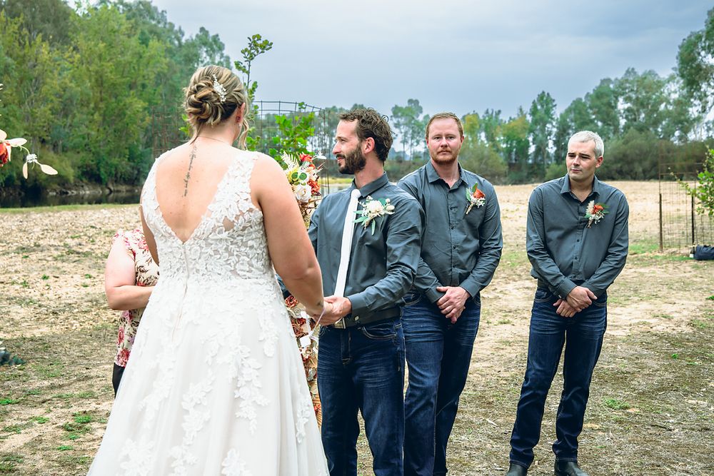 A wedding party stands together outdoors on a sunny day with trees and natural scenery in the background.