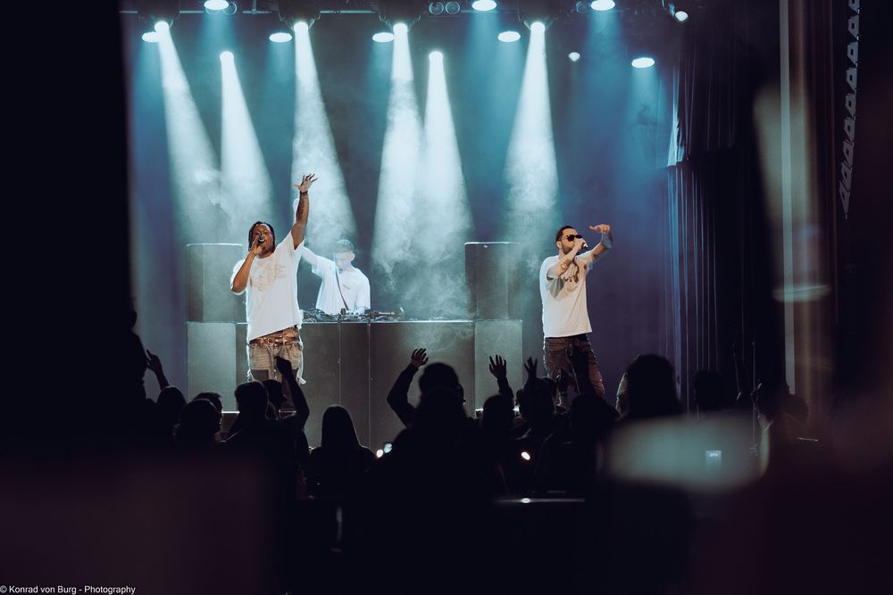 Concert stage with blue spotlights and performers in silhouette against bright background.