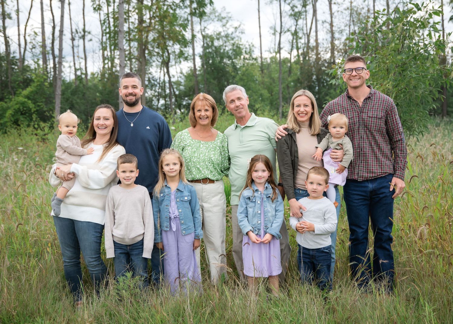 A multi-generational group stands together outdoors in a field with tall grass and trees in the background.