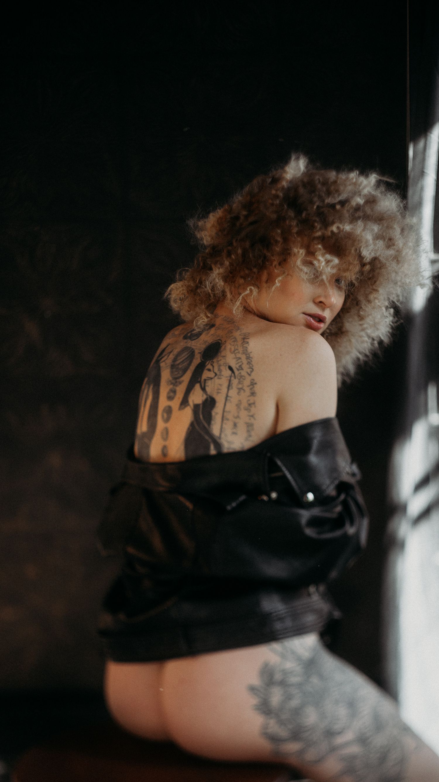 Artistic boudoir photo showing curly haired woman, half nude with tattoos in dramatic lighting against dark background.