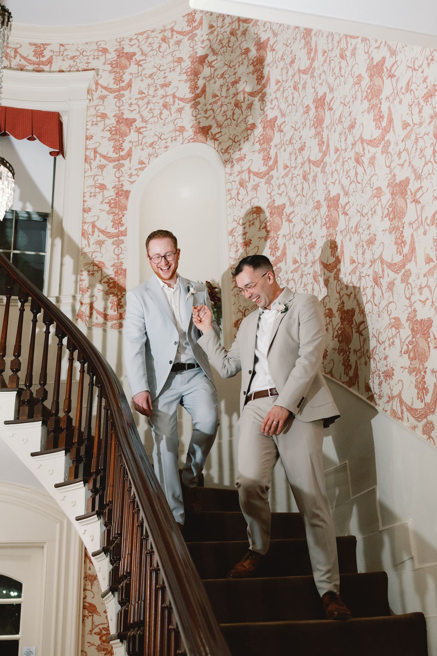 Two people in light suits share a laugh while descending an elegant staircase with floral wallpaper.
