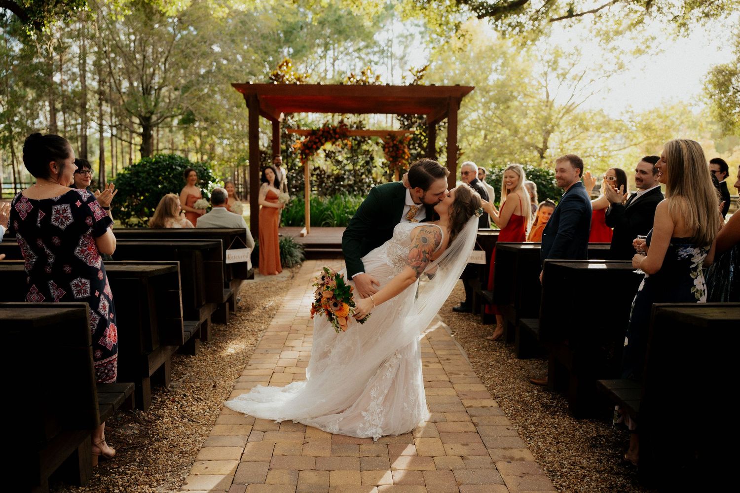 A wedding ceremony takes place on a rustic brick path under a wooden arch surrounded by trees at golden hour at club lake wedding venue