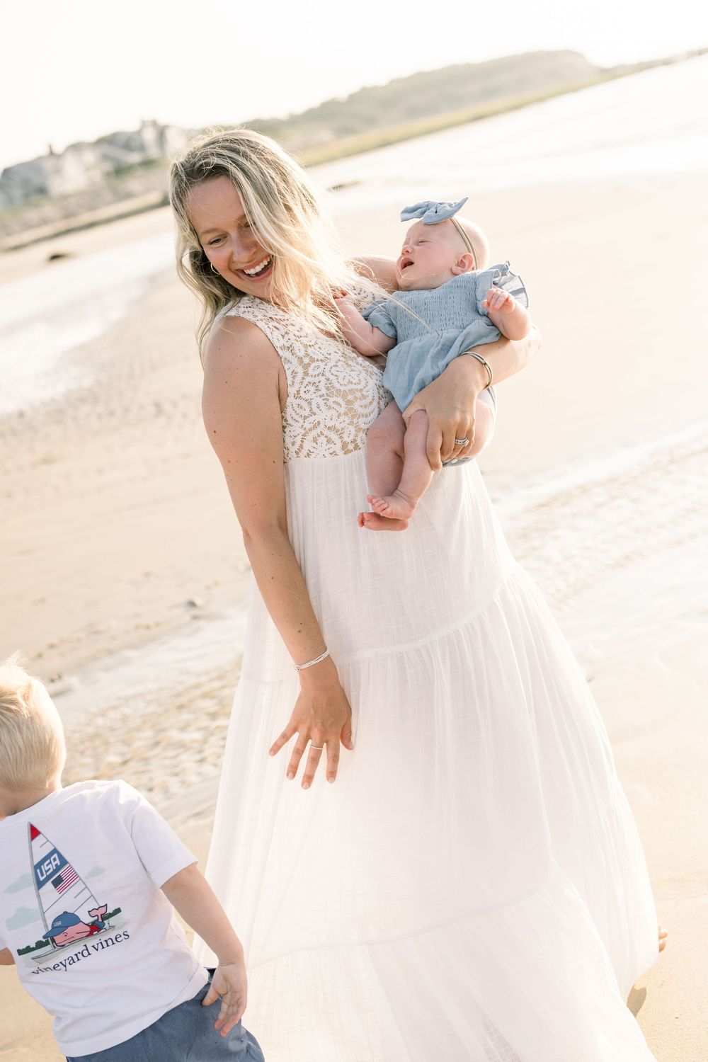 Family in white outfits sharing tender moments together on sunny beach at sunset.