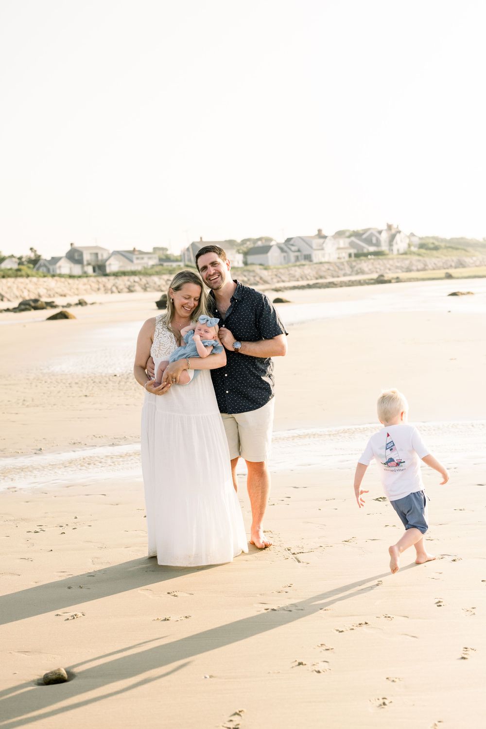 A family walks along a sunny beach shoreline casting long shadows in the sand during golden hour.