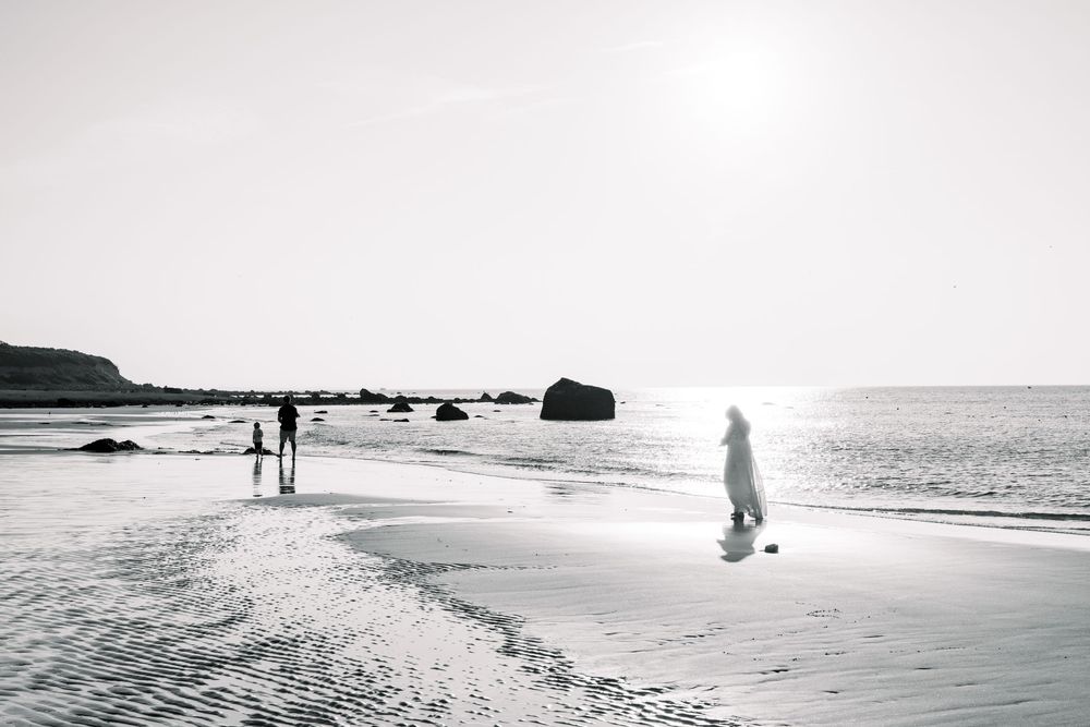Silhouetted figures walk along a misty beach shoreline at sunset with waves crashing against rocks in the distance.
