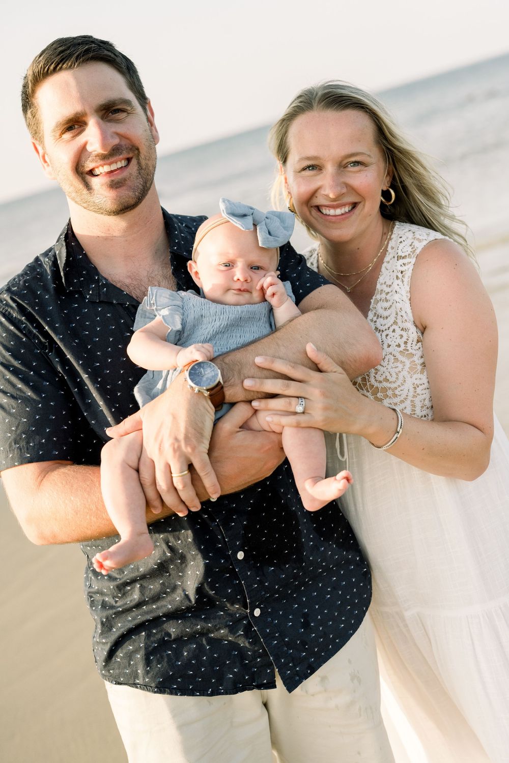A happy family poses together on a beach, with a baby wearing blue against a bright outdoor background.