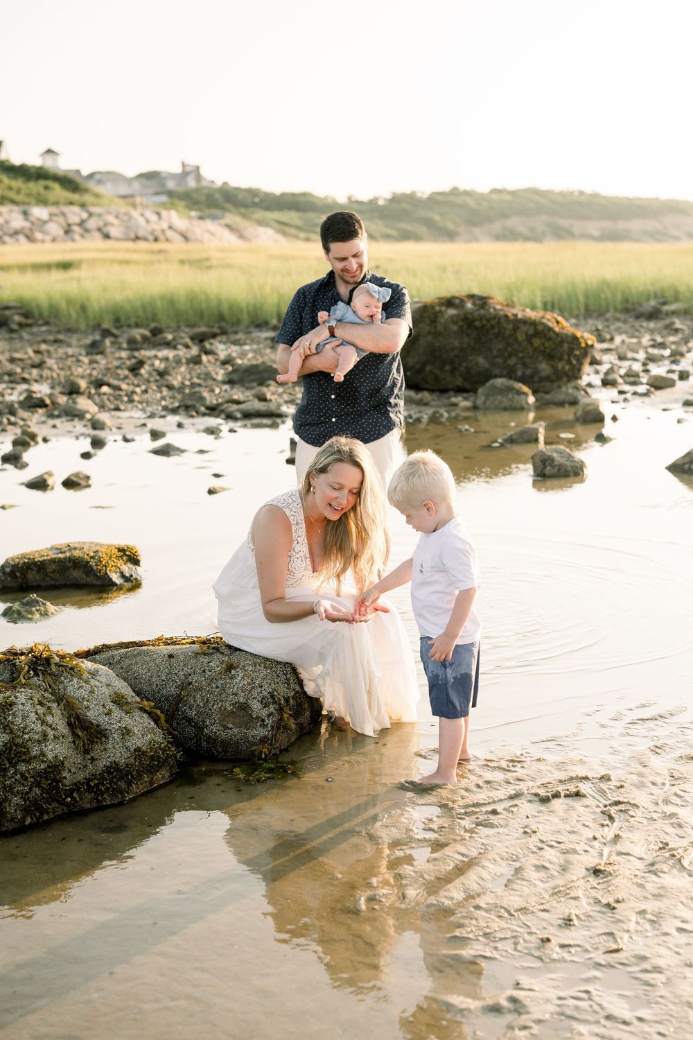 Family exploring tide pools and playing together on a sandy beach at sunset with large rocks and shallow water.