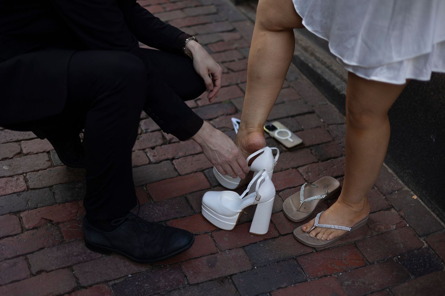 A person helps adjust a white platform heel shoe on a brick sidewalk next to someone wearing sandals.