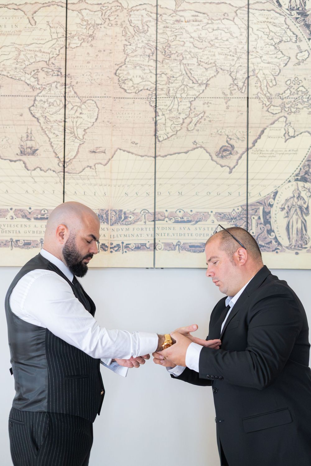 Two men in formal attire stand in front of a vintage world map while exchanging rings during a wedding ceremony.