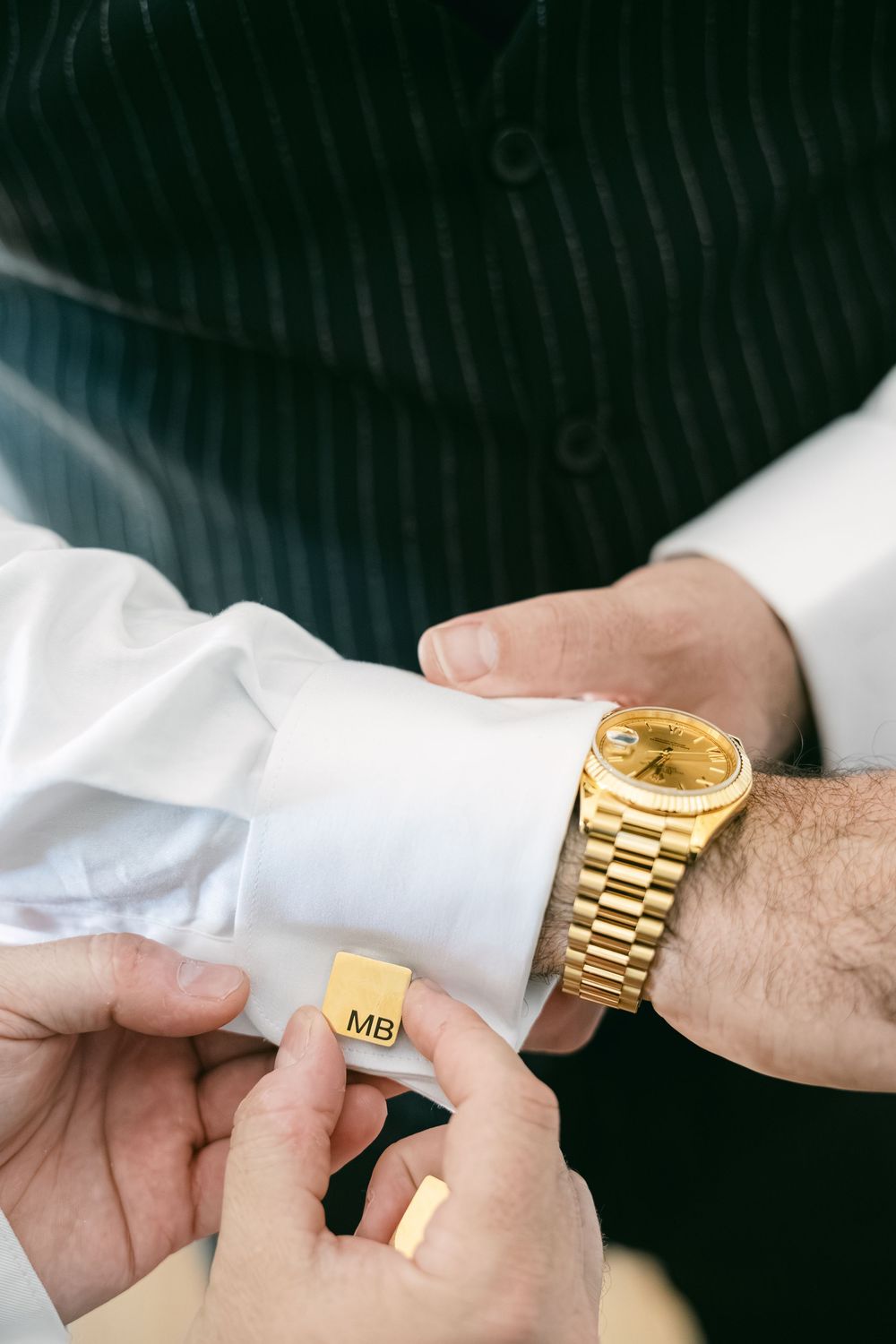 Close-up of hands adjusting gold monogrammed cufflink on white dress shirt sleeve next to luxury gold watch.