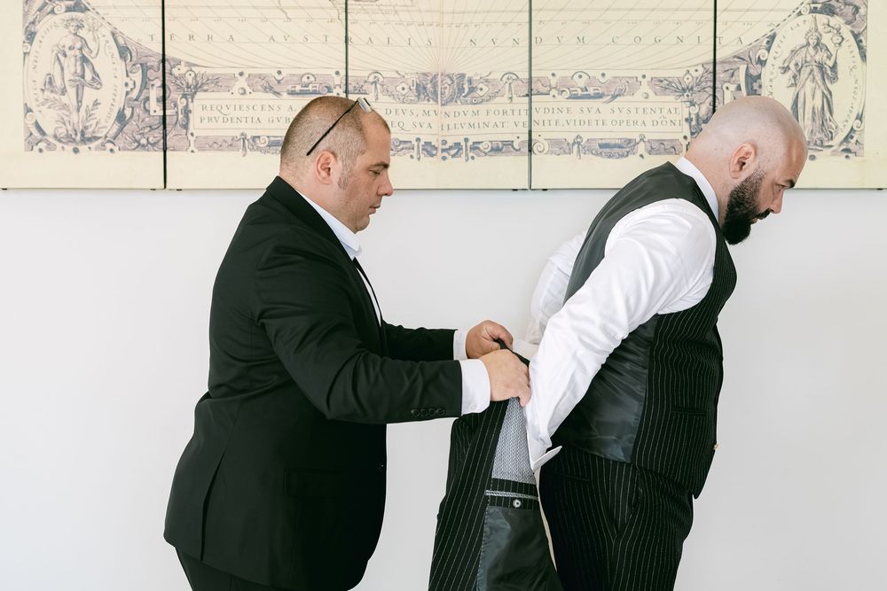 A groomsman helps adjust another's formal attire against a light colored wall with decorative artwork.