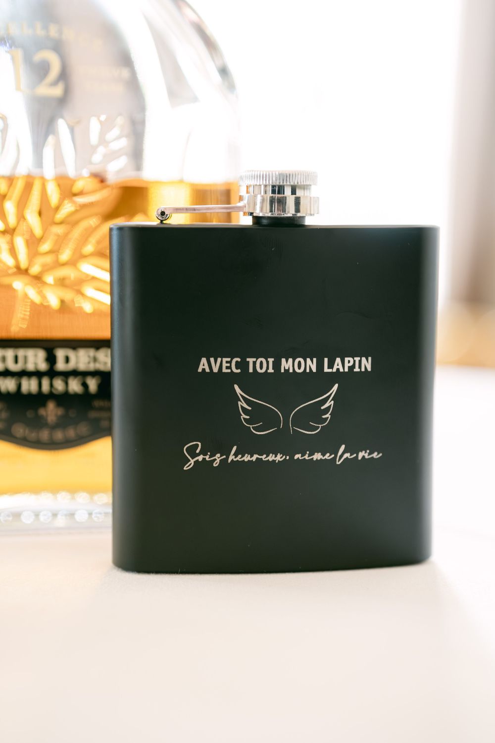 A matte black hip flask with French text 'Avec Toi Mon Lapin' and butterfly design sits on a white surface.