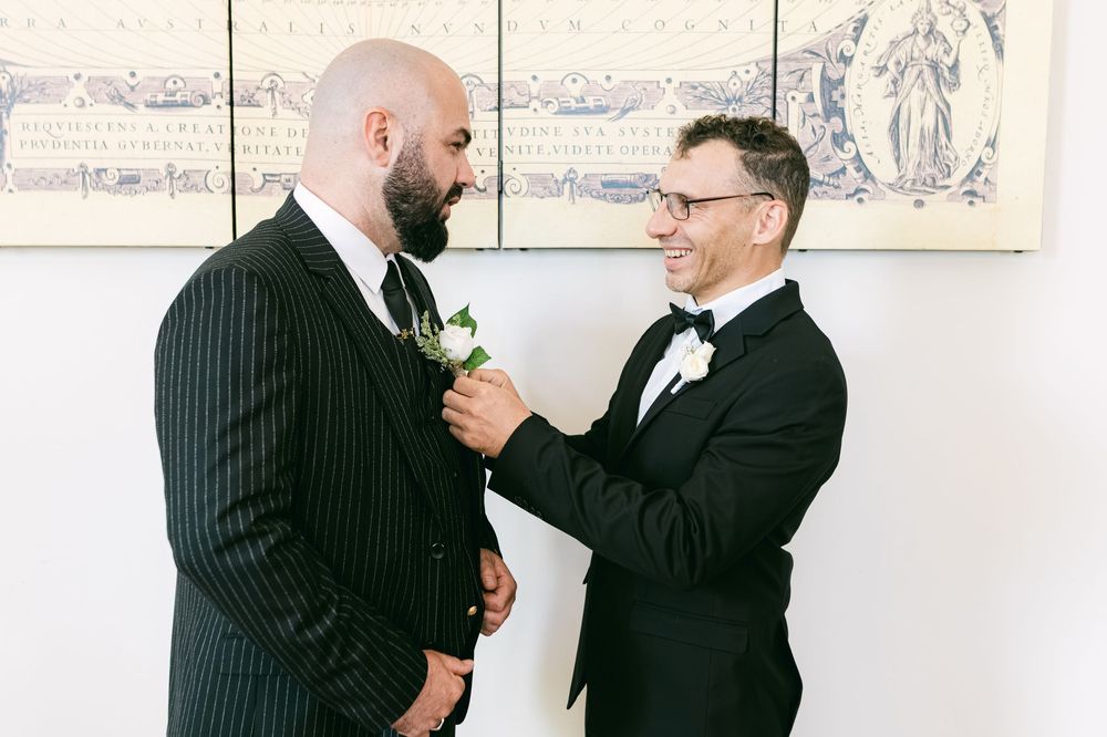 Two people in formal black suits prepare for a special occasion, with one adjusting the other's boutonniere.