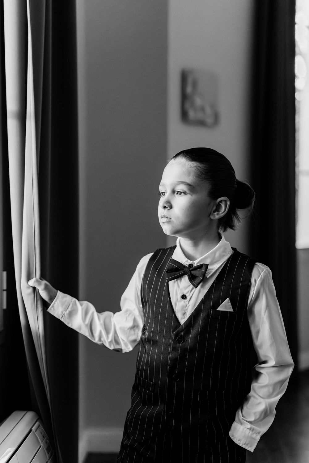 A formal black and white portrait of someone in a pinstriped vest and bowtie looking pensively out a window.