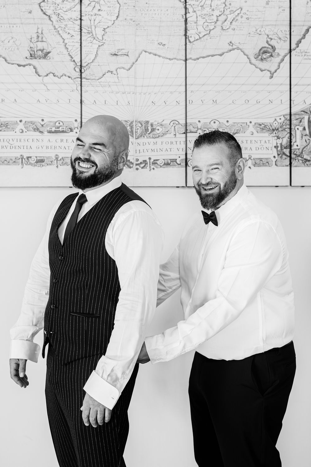 Black and white photo series showing two men in formal attire laughing and posing against a wall with maps.