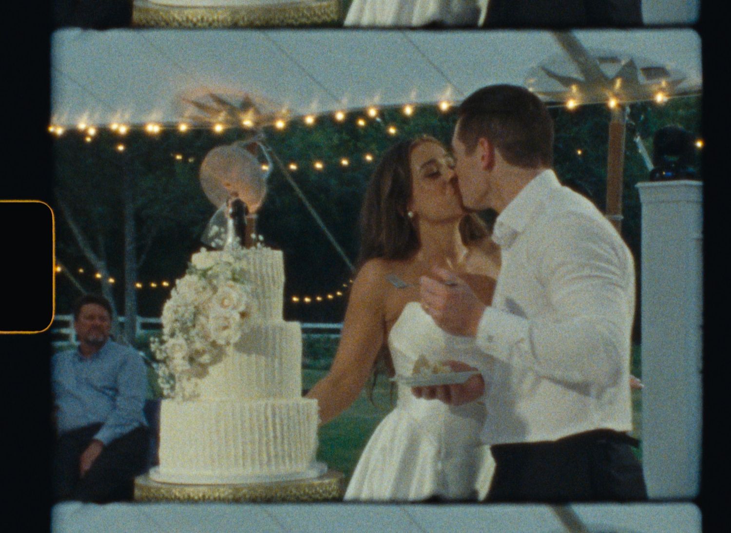 A couple shares a romantic moment next to their elegant white wedding cake under twinkling string lights at an outdoor reception.