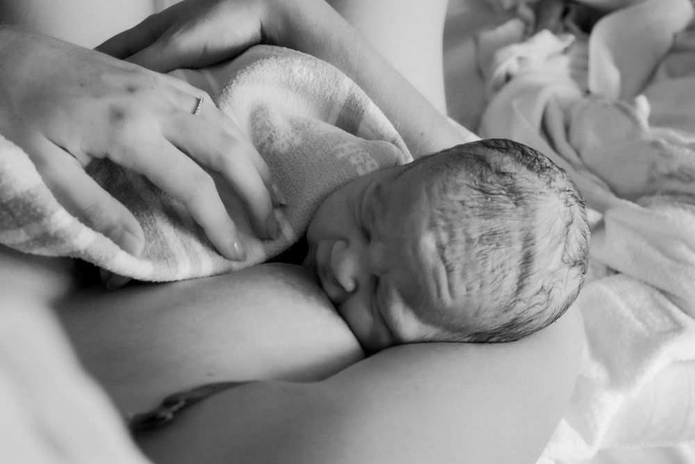 A newborn lies swaddled in white blankets while suckling on their mothers breast in a hospital setting during a central coast birth session.