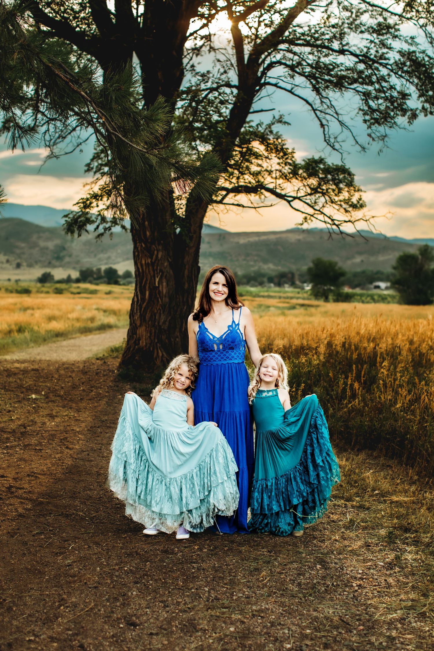 A group wearing coordinating blue and turquoise flowing dresses sits together under a large tree at sunset.