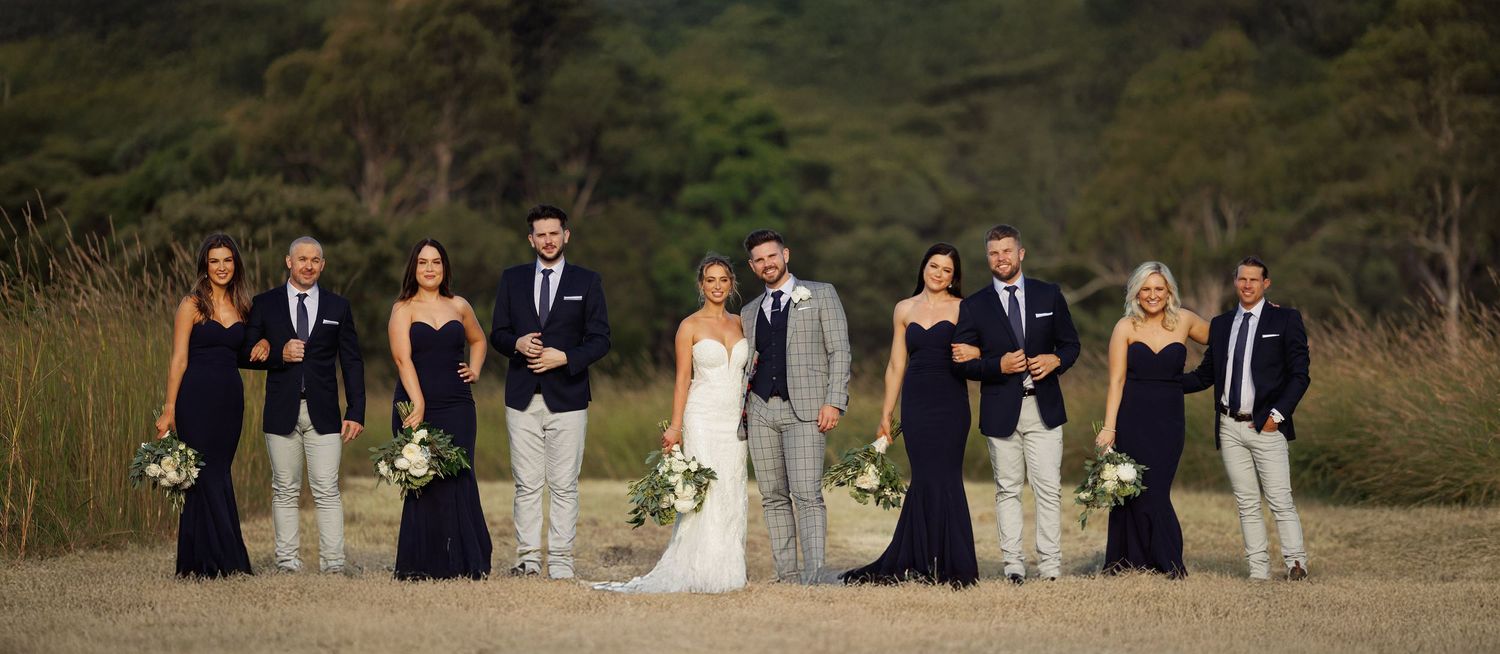 Wedding party of eight posing in navy and grey attire in an open field with lush green hills behind.