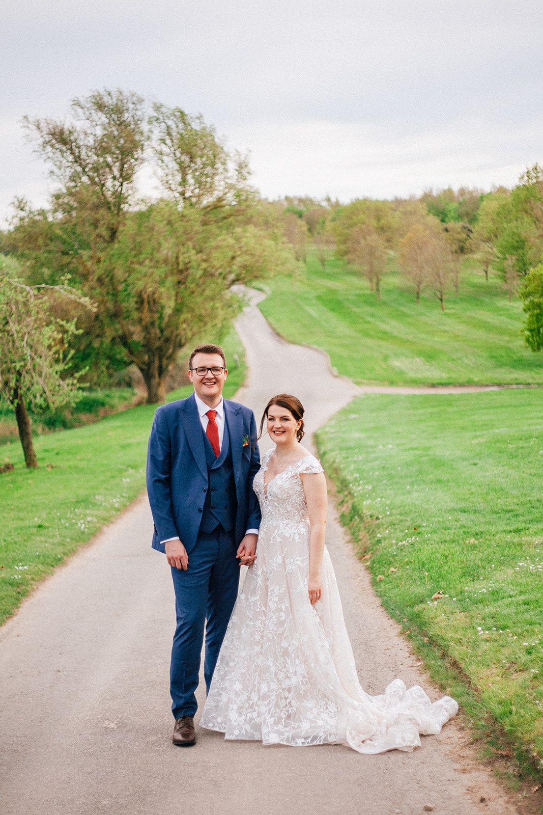 Crondon Park Bride & Groom walks hand in hand down a winding path surrounded by green rolling hills and trees on their wedding day.