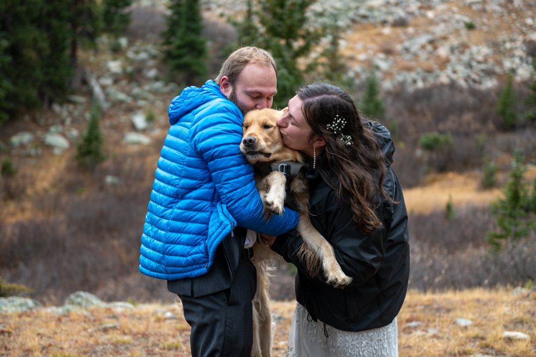 A groom in a blue puffer jacket and black pants and a bride in a white dress and black jacket stand holding their dog and kissing his face.