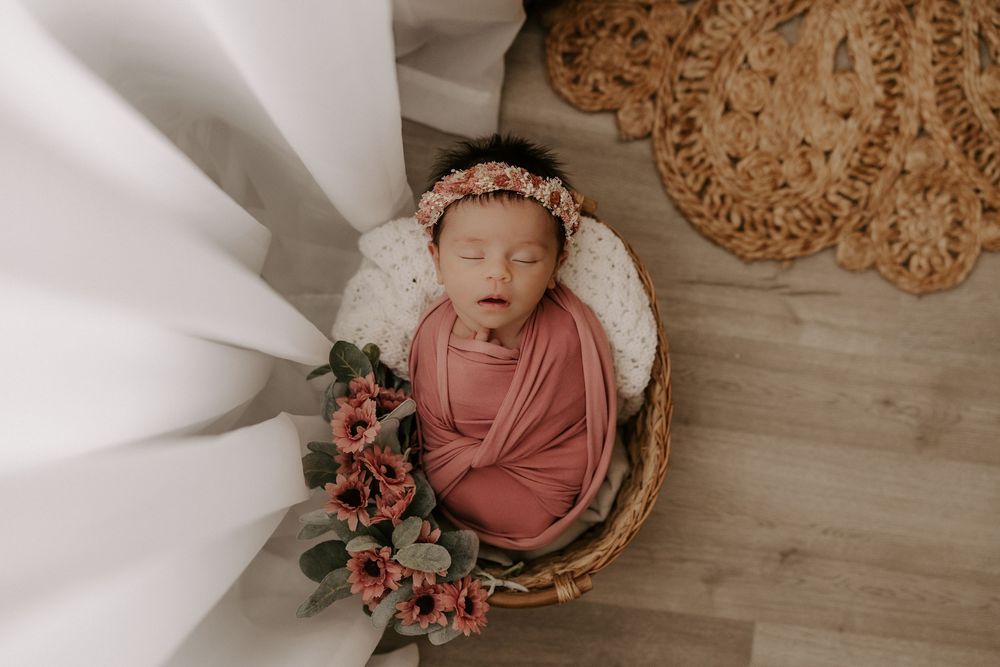 Newborn photography setup with pink swaddle, wooden basket, and delicate floral decorations on light wood floor.