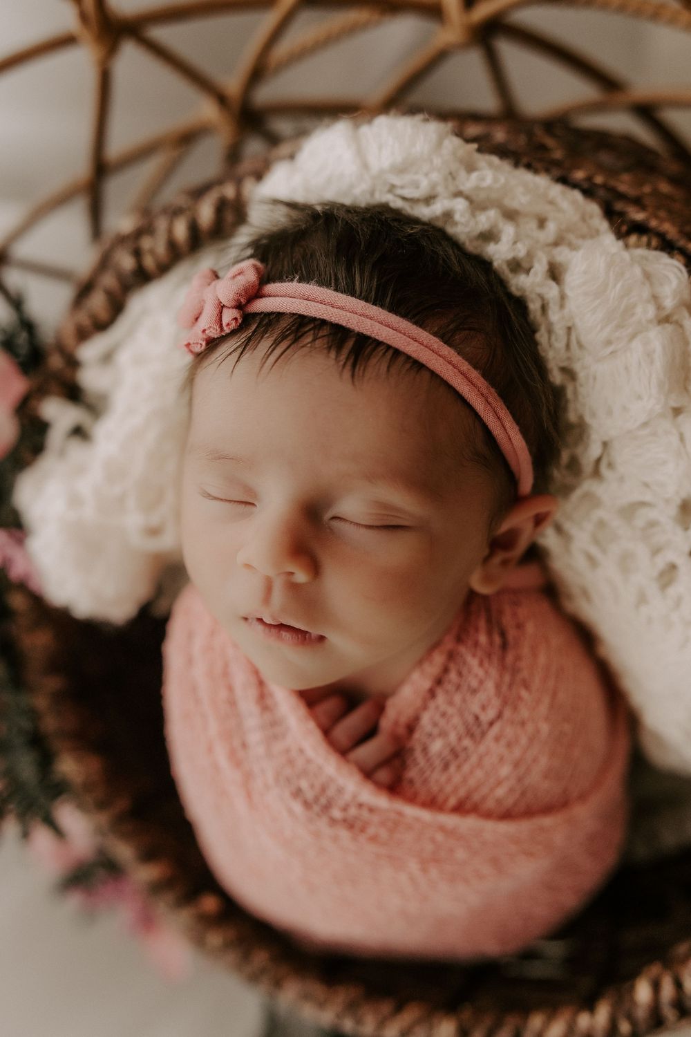 A sleeping newborn baby wrapped in soft pink knit fabric lies peacefully in a cozy basket with white blankets.