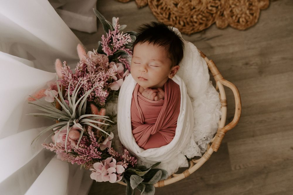 Newborn baby sleeping peacefully in white bowl with pink floral arrangement on wooden floor.